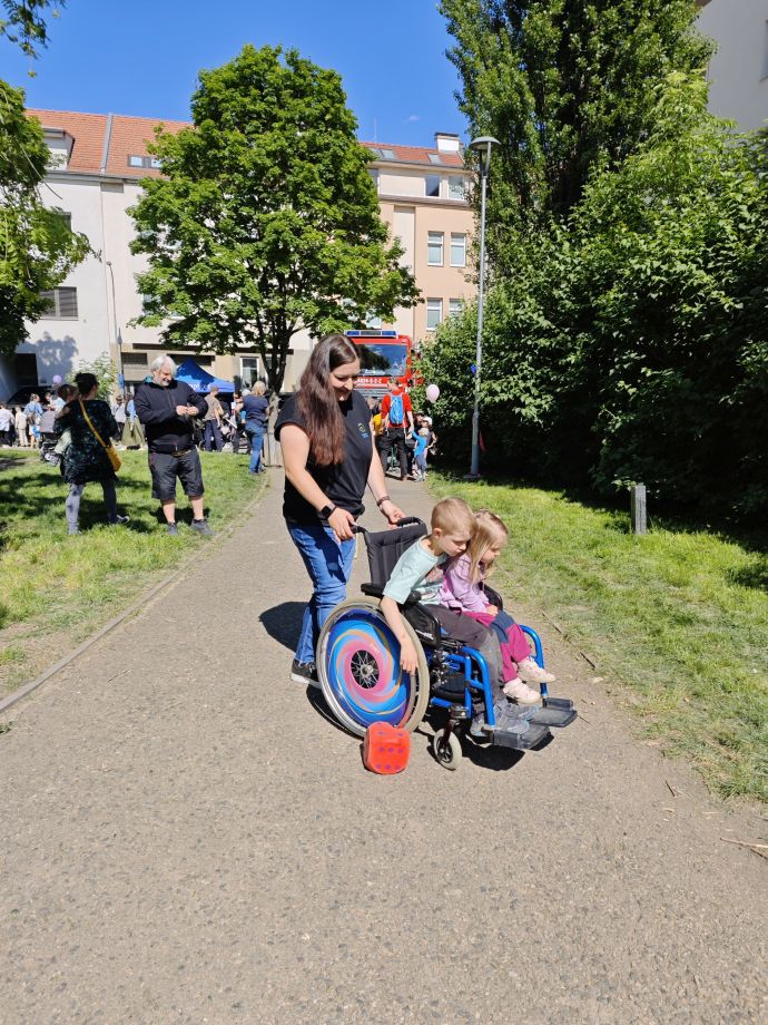 Dvě malé děti sedí na invalidním vozíku a za pomoci dospělé ženy zkouší slalom na vozíku